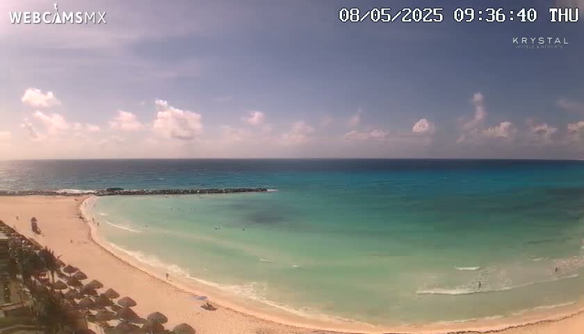 A partly cloudy sunny day shows a beach with calm, clear turquoise water and light sand, with some people on the beach and in the water.