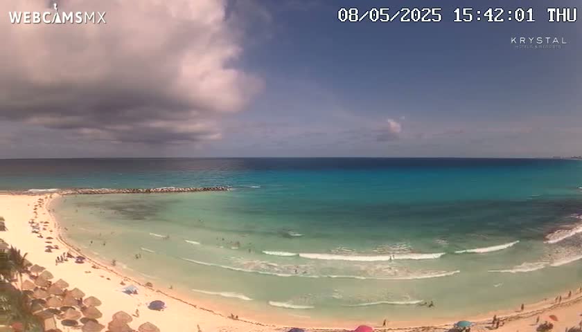 A partly cloudy sky overlooks a beach with turquoise water and gentle waves, where people are swimming and relaxing under beach umbrellas.