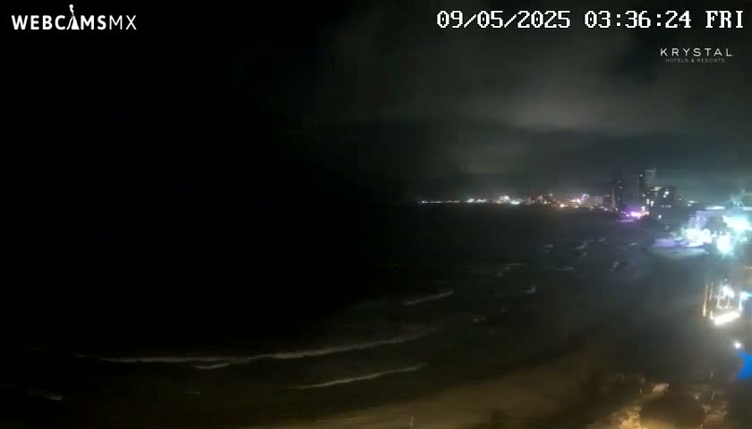 A nighttime coastal scene shows dark, wavy water lapping a beach under a cloudy sky.