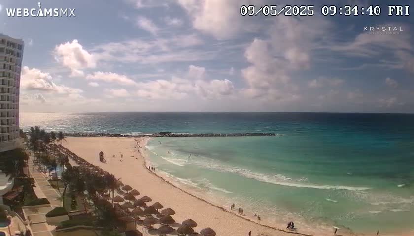 A partly cloudy day reveals a sandy beach with turquoise water, gently lapping at the shore, near a high-rise hotel.