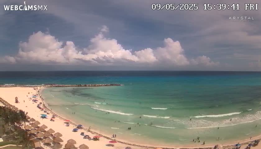 A partly cloudy day shows a beach with light waves, turquoise water, and many people swimming and relaxing under beach umbrellas.