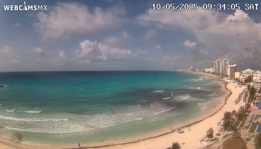 A partly cloudy sky overlooks a long sandy beach with turquoise water lapping gently at the shore, with hotels visible in the distance.