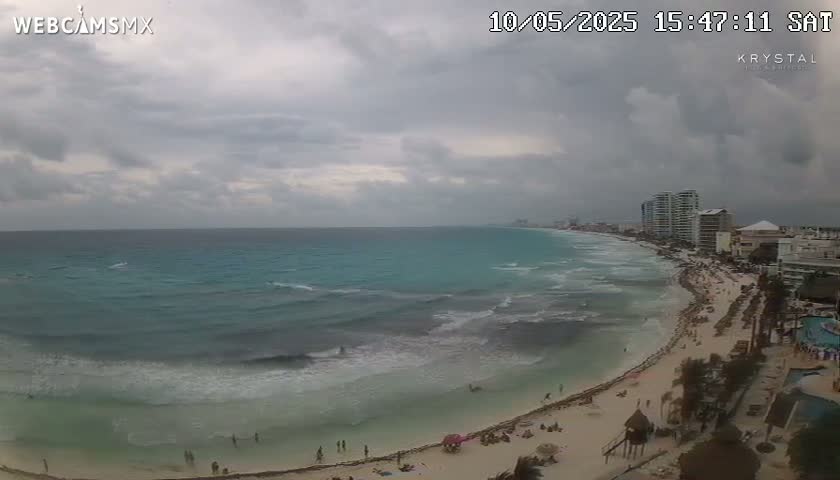 A beach with turquoise water and moderate waves is seen under an overcast sky, with buildings and people visible along the shoreline.