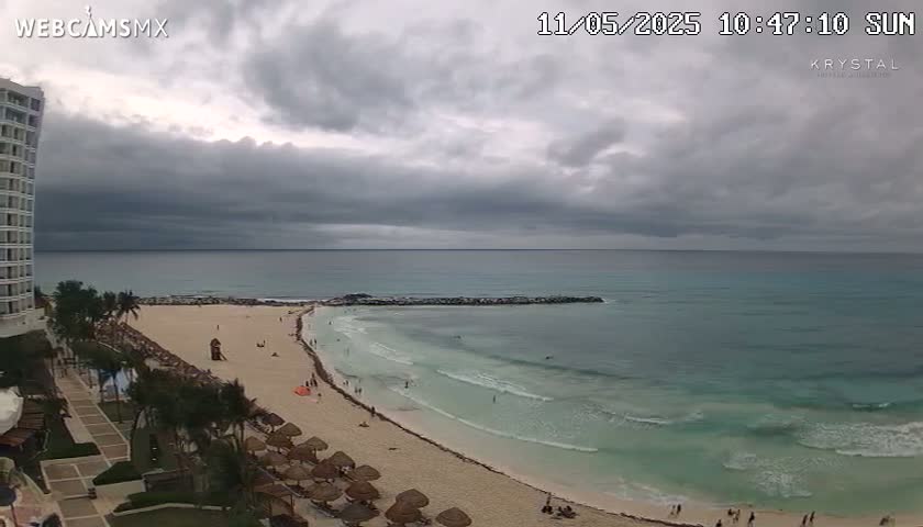 A sandy beach with turquoise water and a few people is seen from a high vantage point overlooking a resort on an overcast day.
