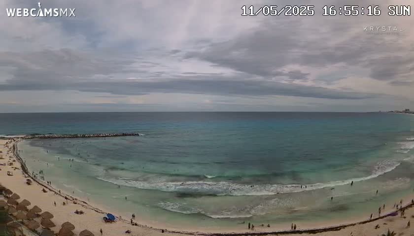 A mostly cloudy day shows a sandy beach with turquoise water and small waves, sparsely populated with people.