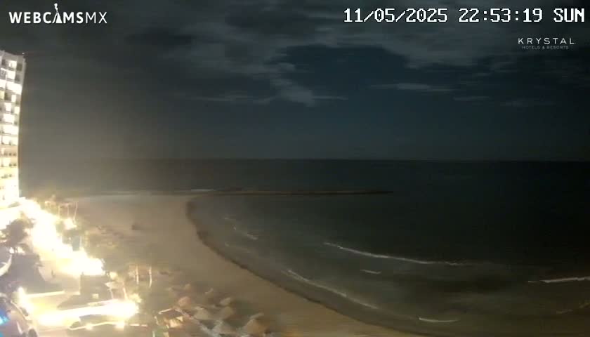 A nighttime view of a beach and ocean under a mostly cloudy sky, with a brightly lit building visible on the left.