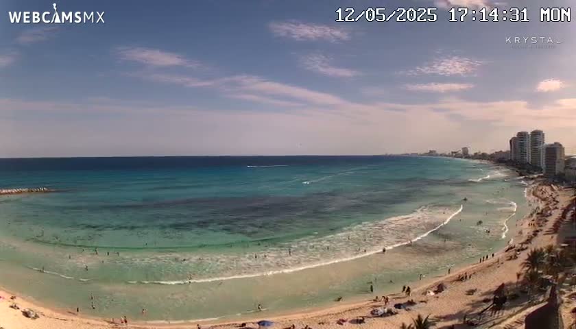 A mostly sunny day at a beach with clear, shallow turquoise water lapping a sandy shore, with high-rise buildings visible in the distance.