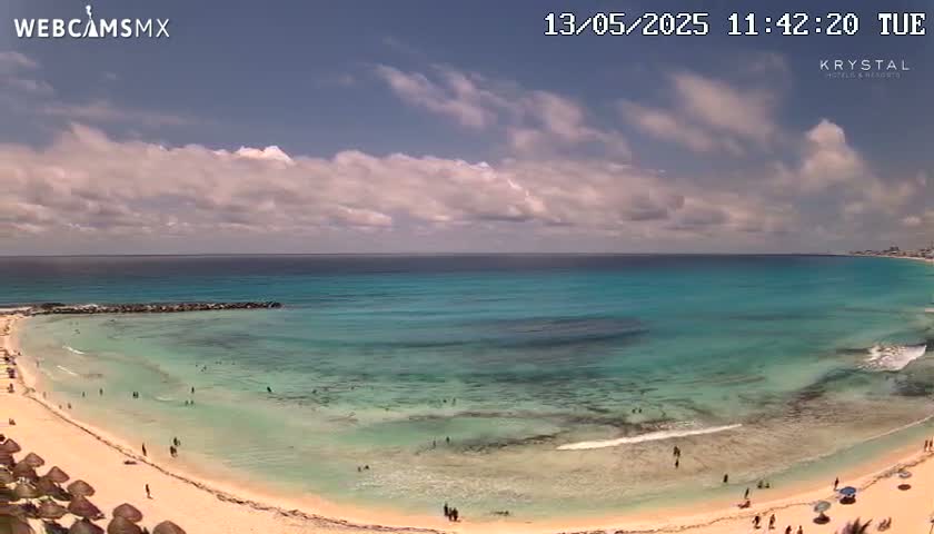 A partly cloudy day reveals a calm, turquoise ocean lapping a sandy beach with scattered beachgoers.