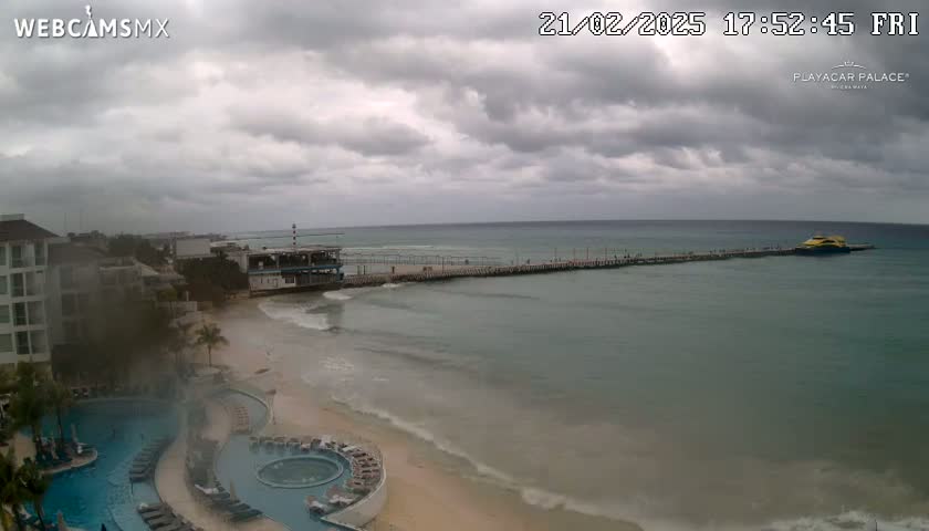 A resort with pools overlooks a beach and pier with a boat docked under an overcast sky.