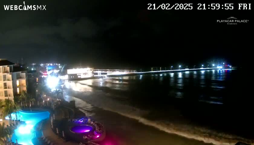 A nighttime beach scene shows a brightly lit pier extending into dark water with gentle waves lapping the shore near a resort with illuminated pools.