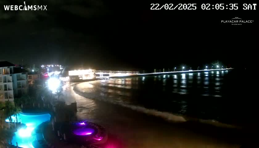 A nighttime beach scene shows a brightly lit pier extending into dark water with gentle waves lapping the shore near a resort with illuminated pools.