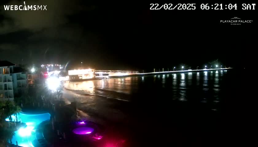A nighttime view of a beach with a brightly lit pier extending into the dark ocean, illuminated pools, and a building visible in the background under a cloudy sky.