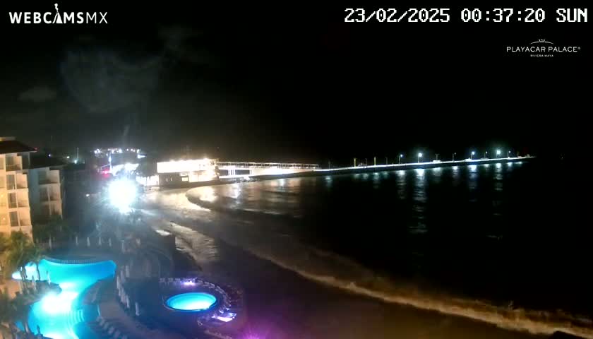 A nighttime beach scene shows a resort with illuminated pools and a pier extending into gently lapping waves under a dark sky.