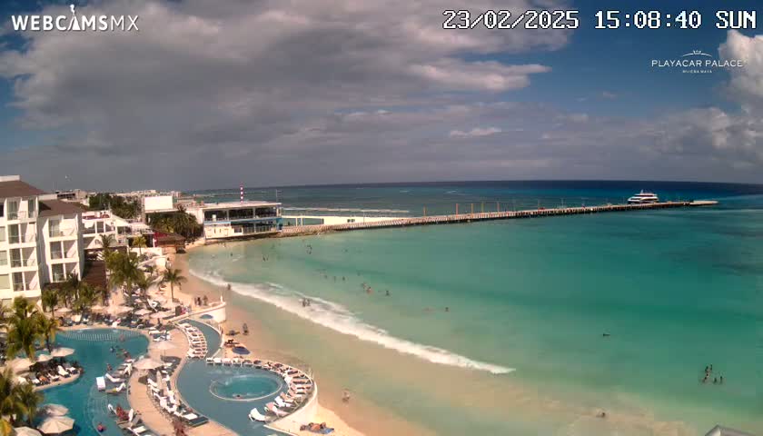 A partly cloudy sunny day shows a beach resort with turquoise water, a long pier with a boat, and people swimming and sunbathing.