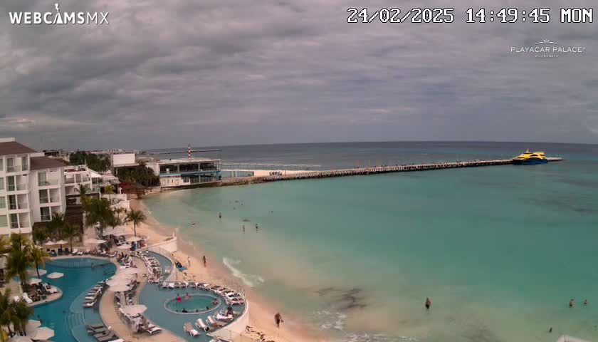 An overcast day shows a resort with a pool area next to a beach and pier with a boat, all overlooking turquoise water.