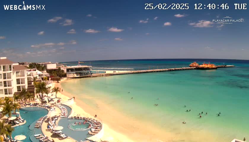 A beach resort with multiple pools overlooks a calm, turquoise ocean with a pier and two boats moored; the sky is mostly sunny with a few scattered clouds.