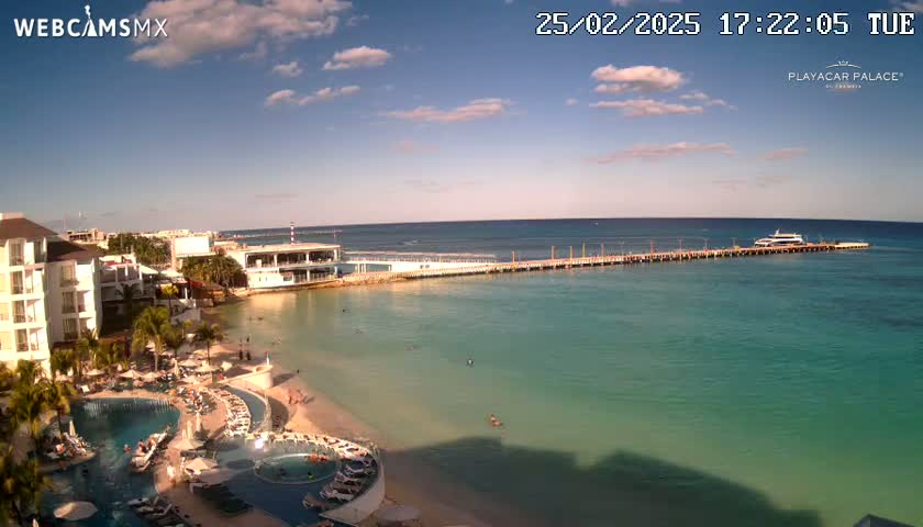 A resort beach scene shows a long pier extending into clear, teal water under a partly sunny sky.