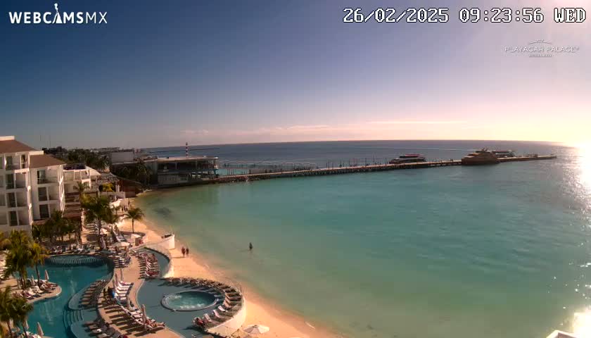 A resort with pools and a sandy beach overlooks a calm, turquoise ocean with a pier and several boats under a clear, sunny sky.