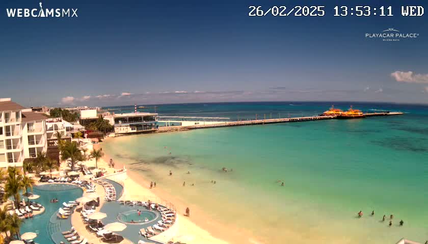 A resort with multiple pools overlooks a clear, turquoise ocean where several people are swimming, with a pier and two boats visible in the distance under a sunny sky.