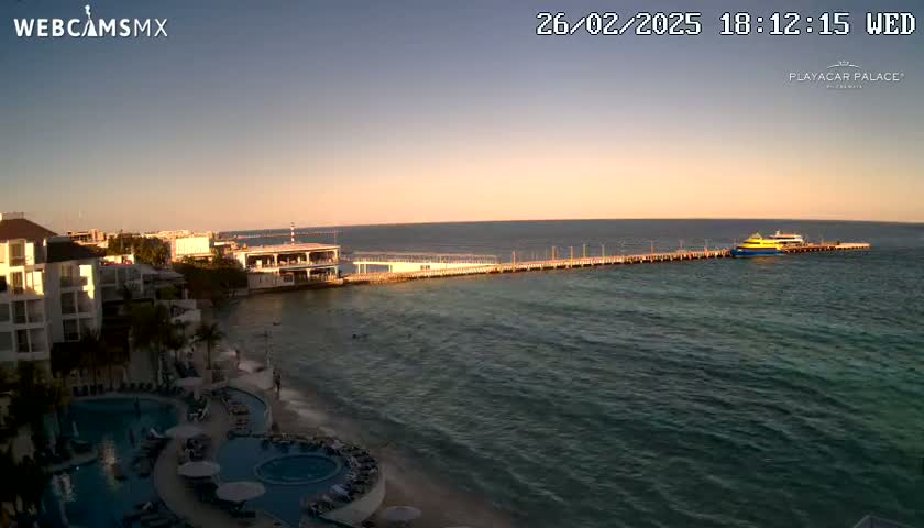 A resort with pools sits on a beach next to a pier with a ferry docked at sunset under a clear sky.