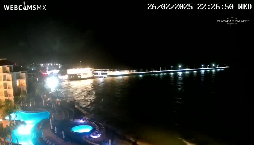 A nighttime view of a resort with illuminated pools next to a dark ocean with a brightly lit pier extending into the water.