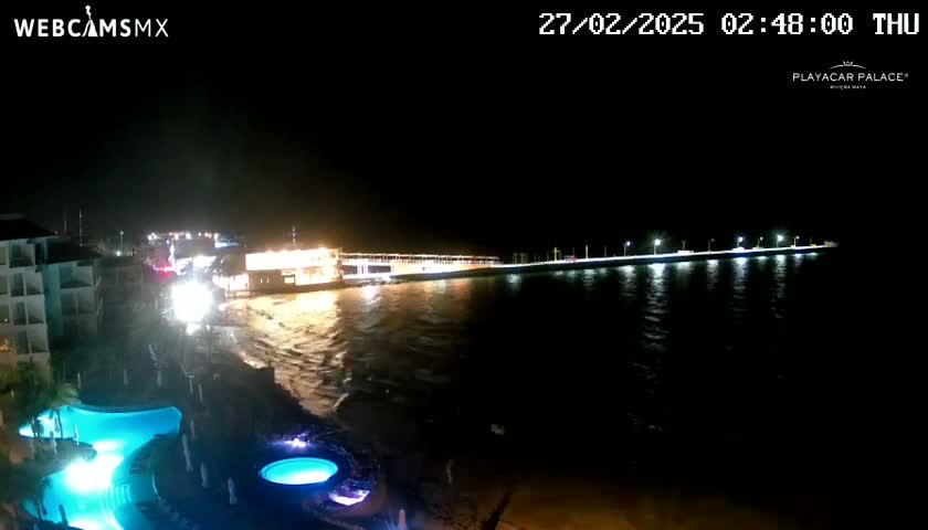 A brightly lit pier extends over dark, wavy water at night, adjacent to a resort with illuminated pools.