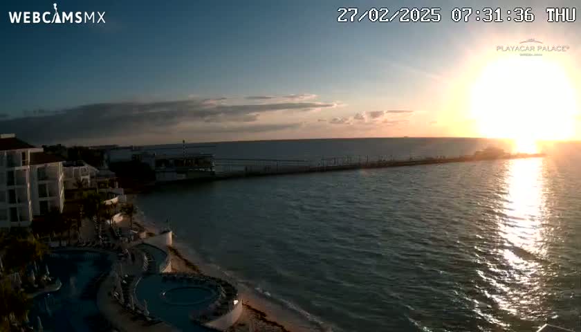 A sunrise over calm ocean water illuminates a resort with pools and a pier extending into the water.