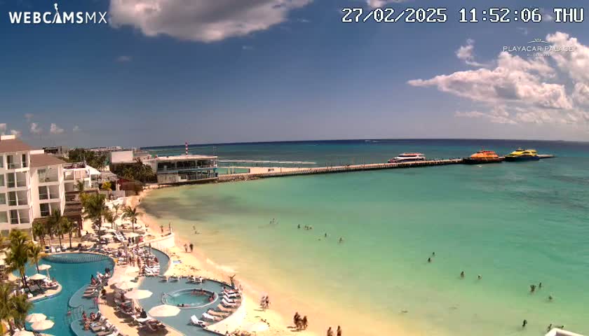 A partly cloudy sunny day at a beach resort shows a white building, multiple pools, a sandy beach, turquoise water, and several boats docked at a pier.