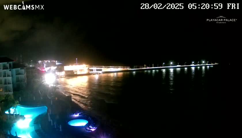 A nighttime coastal scene shows a resort with illuminated pools, a pier with lights, and dark, wavy water under a cloudy sky.