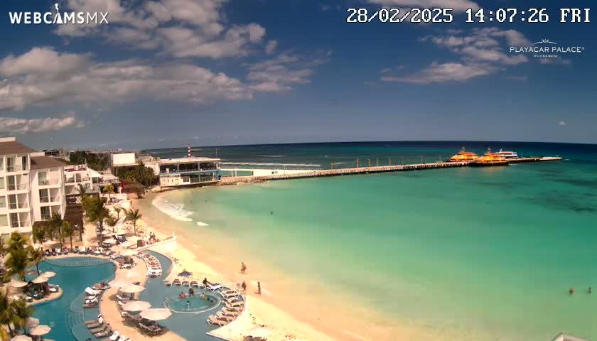 A beach resort with turquoise water, a sandy beach, swimming pools, and several boats docked at a pier under a partly cloudy sky.