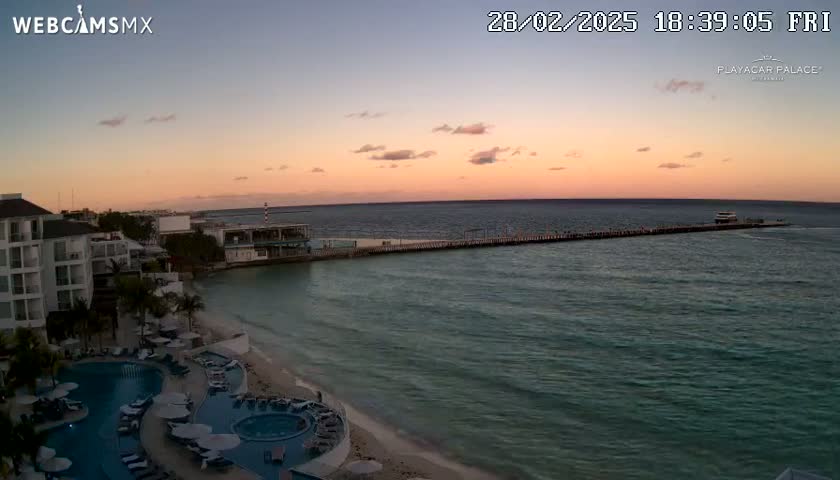 A coastal resort with pools at sunset, overlooking a calm ocean with a pier extending into the water.