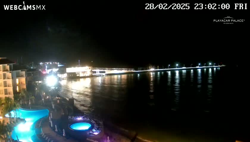 A nighttime view of a resort area shows a lit-up waterfront with a long pier extending into calm water, and a resort with pools illuminated in blue.