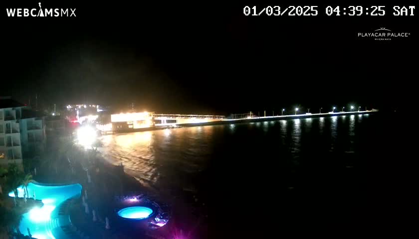 A nighttime view of a resort area with illuminated pools and a brightly lit pier extending into dark water.