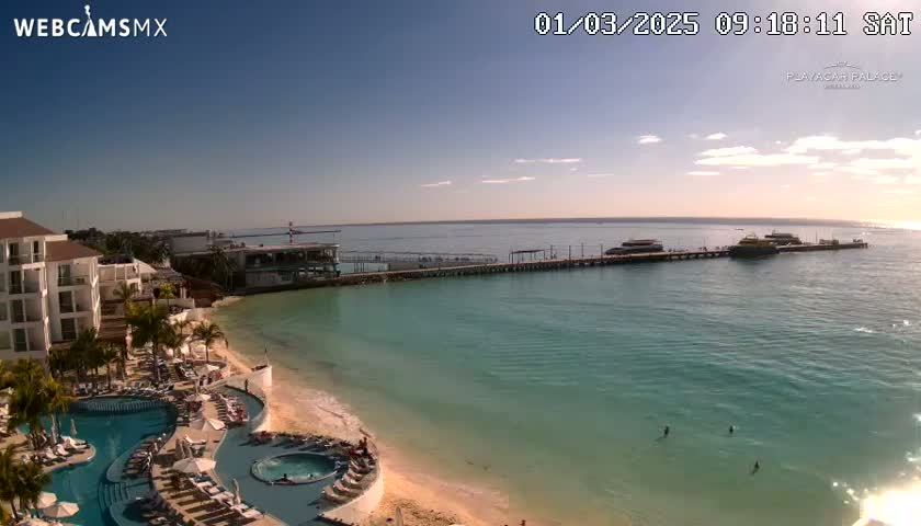 A beachfront resort with a swimming pool overlooks a long pier with boats moored, under a mostly clear, sunny sky.