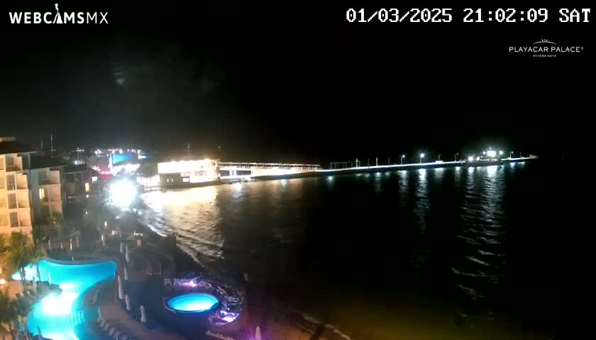A nighttime view of a resort area with a long pier extending into a calm body of water, illuminated by lights, and featuring pools in the foreground.