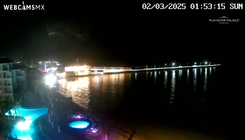 A nighttime view of a resort area with illuminated buildings and a pier extending over dark water, under a mostly clear sky.