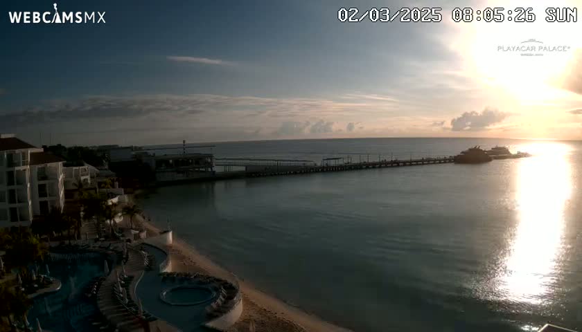 A sunrise over calm ocean water illuminates a long pier with boats at its end, next to a resort with pools and a beach.