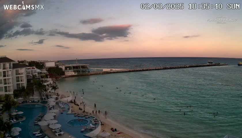 A beach resort at sunset, with calm turquoise water and a pier extending into the sea, under a partly cloudy sky.