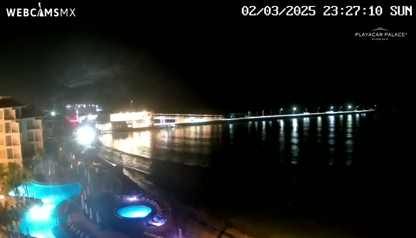A nighttime coastal scene shows a brightly lit pier extending into dark water, with a building and swimming pools visible in the foreground under a clear, dark sky.