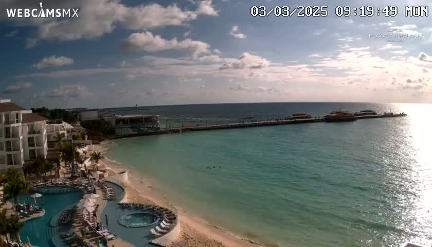 A partly sunny day shows a resort with a pool overlooking a beach and calm turquoise water, with a pier and boats in the distance.
