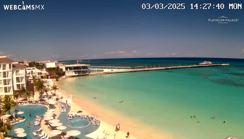 A sunny day at a beach resort shows a long pier extending into clear turquoise water, with a boat docked at the end, and a sandy beach with a swimming pool and resort buildings visible.