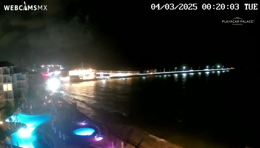 A nighttime view of a resort with illuminated pools, a beach, and a pier extending into dark, wavy water under a cloudy sky.