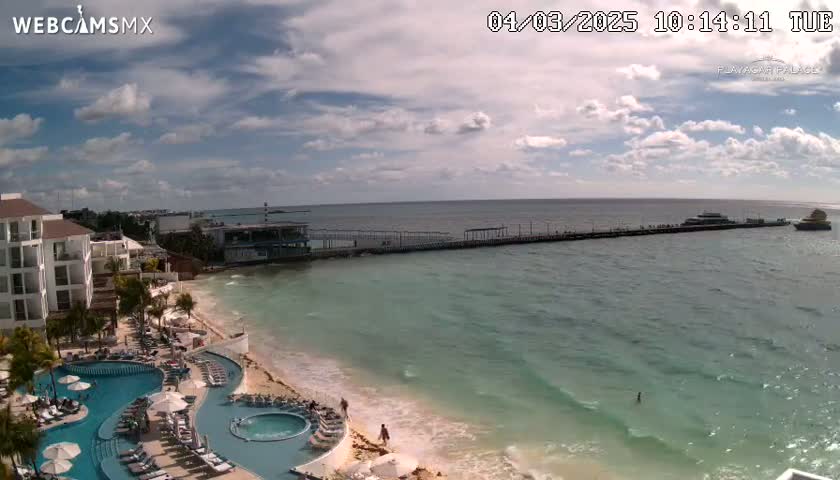 A partly cloudy day shows a resort with multiple pools overlooking a calm, turquoise ocean and a long pier with boats.