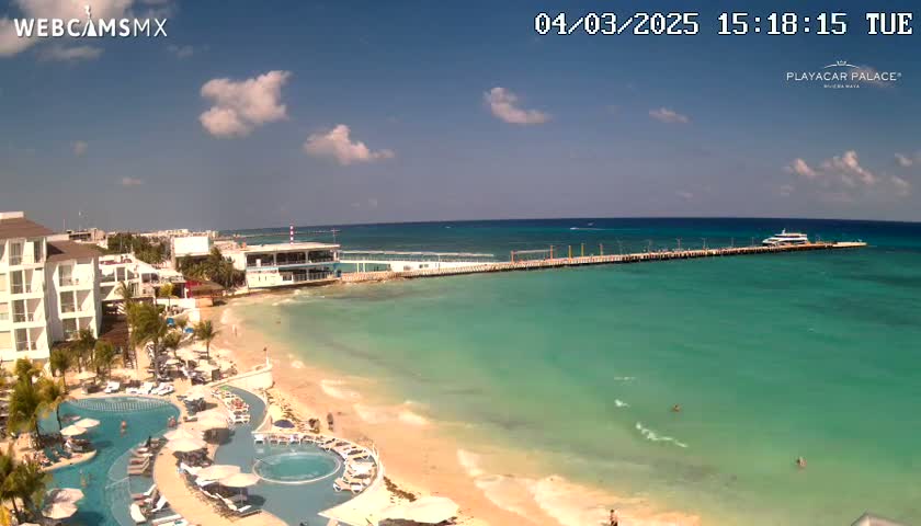 A resort with multiple pools sits on a beach next to a long pier with a boat docked at the end, all under a partly cloudy sky.
