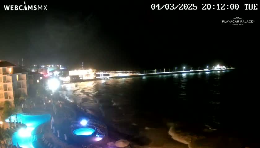 A nighttime view of a resort with illuminated pools near a shoreline with moderately active waves under a dark sky.