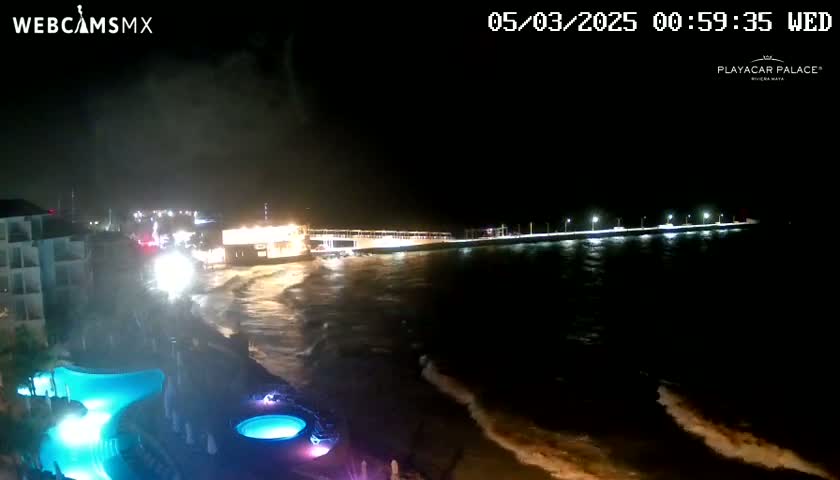 A nighttime coastal scene shows a resort with illuminated pools near a pier extending into a dark ocean with moderate waves under a cloudy sky.