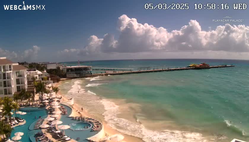 A resort with pools overlooks a beach with turquoise water and waves, under a partly cloudy sky.