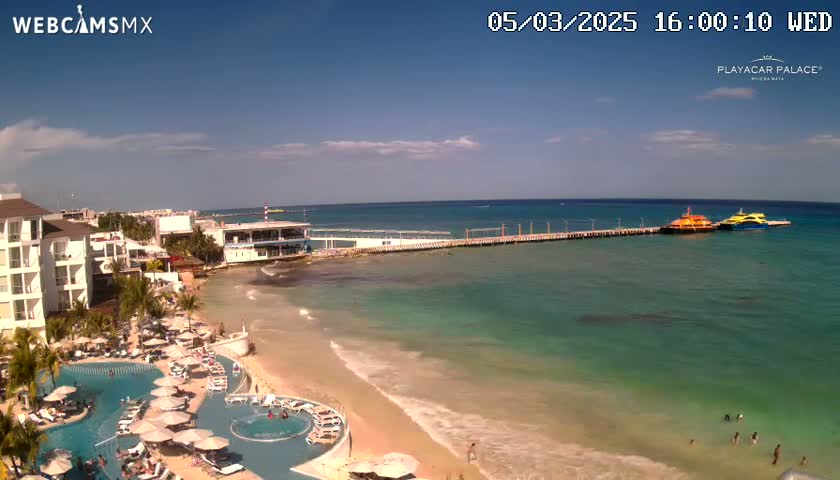 A resort with a swimming pool overlooks a beach with calm, clear turquoise water and two boats docked at a pier under a sunny sky.