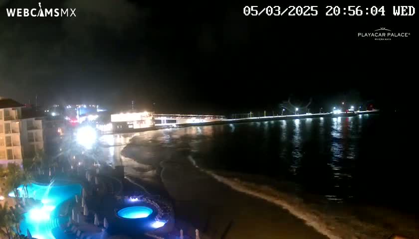 A nighttime view of a resort's illuminated pool area overlooking a calm, dark ocean with a pier in the distance under a clear, dark sky.