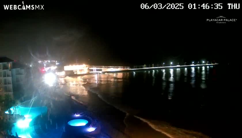 A nighttime view of a beach with a brightly lit pier extending into dark water, illuminated pools are visible in the foreground.